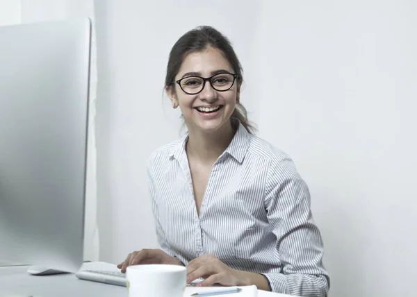 A happy indian girl at the computer, in the workplace. A young woman in glasses rejoices at her achievements.