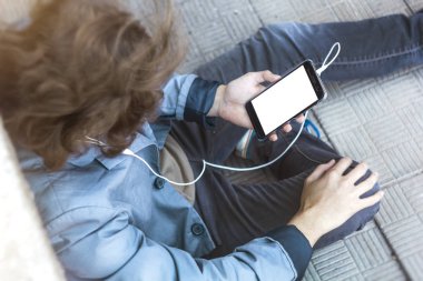 Genç bir adam elinde bir telefon tutuyor ve müzik dinliyor. Genç bir çocuk sokakta otururken bir alet kullanıyor..