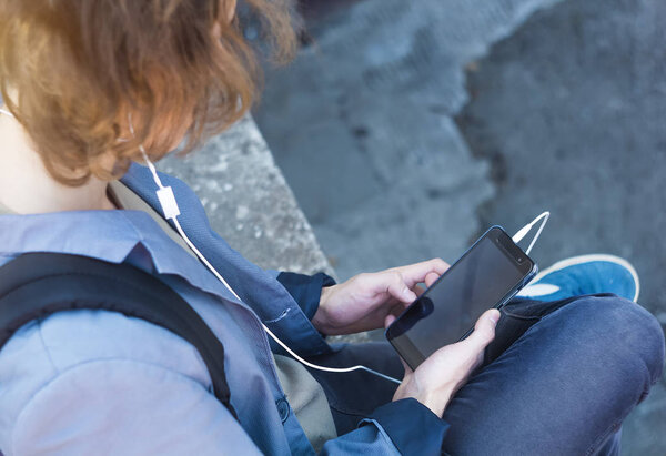 A young man holds a phone in his hand and listens to music. A boy teenager uses a gadget while sitting on the street.