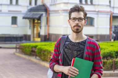 Gözlük takan bir öğrenci adam bir klasör bir üniversite arka plan elinde tutuyor. Şık bir genç adam üniversite standları.