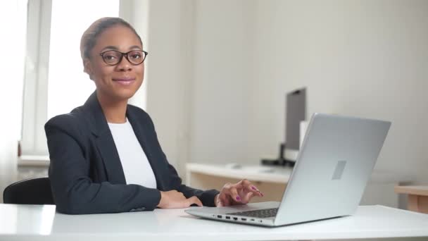 一个穿着西装 面带微笑的非洲裔美国经理在笔记本电脑后面的办公室里工作的画像 友好的黑人女孩 戴着眼镜坐在办公桌前 — 图库视频影像