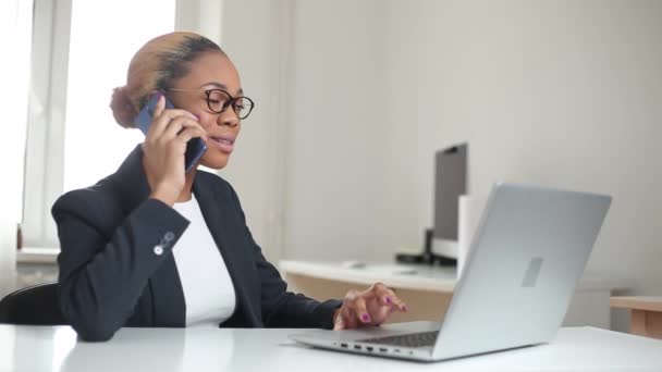ビジネススーツのアフリカ系アメリカ人の若い女性マネージャーが電話で優しい話をしています 成功した女性は コンピュータでオフィスで働き クライアントに助言します — ストック動画