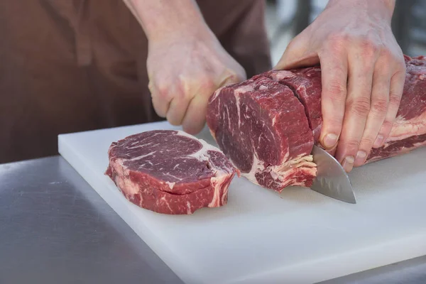 Carniceiro a cortar carne na cozinha. Fecho de mãos com uma faca e carne . Imagens Royalty-Free