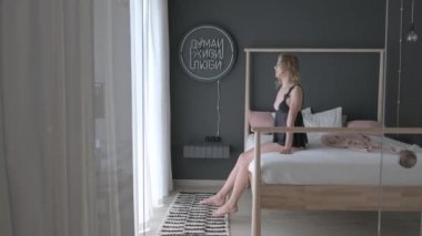 Güzel sarışın yataktan kalkar ve perdeleri çeker. Güneş Büyük Pencereden Üzerine Parlıyor. Mutlu Genç Kız Yeni Günü Kutluyor