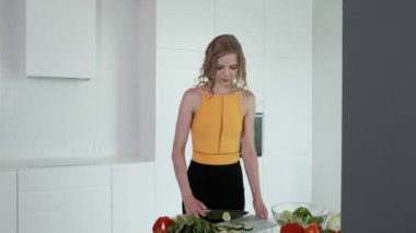 Sebze salatası için salatalık doğrayan genç bir kadın. Beyaz modern mutfak. Sebze, domates, kırmızı biber, marul, salatalık, avokado. 4k