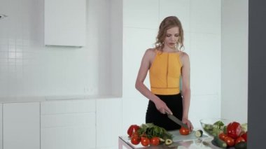 Sebze salatası için domates doğrayan genç bir kadın. Beyaz modern mutfak. Sebze, domates, kırmızı biber, marul, salatalık, avokado. 4k.