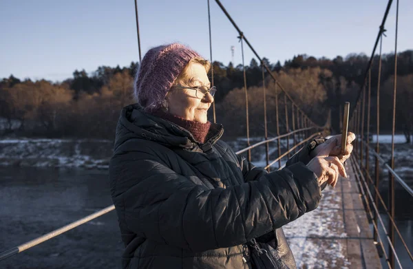 Літня жінка в окулярах фотографується на своєму телефоні, стоячи на підвісному мосту — стокове фото
