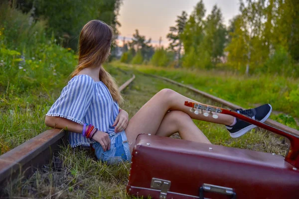 Uma adolescente está sentada nos trilhos com uma guitarra e uma mala velha — Fotografia de Stock