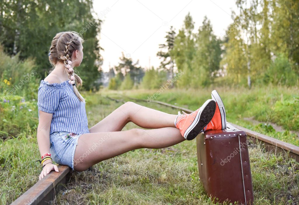 A teenage girl is sitting on the railway tracks with her feet on a large suitcase.
