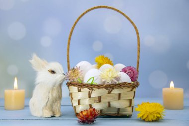 Paskalya Tavşanı, kurumuş çiçeklerle süslenmiş yumurta dolu bir sepetin yanında oturur. Arka planda yanan mumlar.