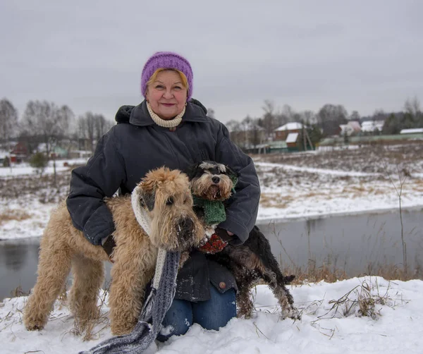 Photo of an elderly woman with two dogs on a snow-covered river Bank against the background of a village.