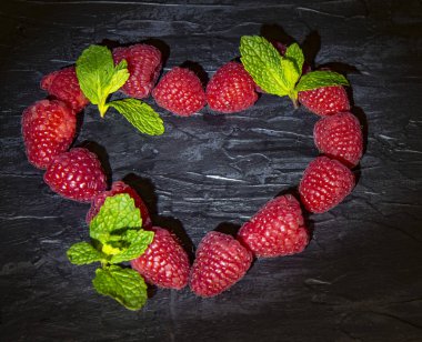 Taze ahududu ve nane yaprakları kara bir zemin üzerinde kalp şeklinde serilmiş..
