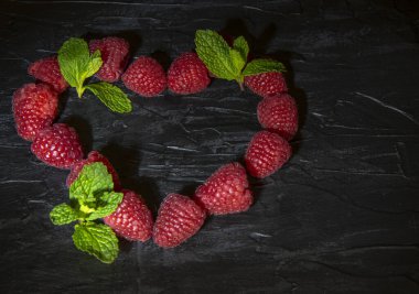 Taze ahududu ve nane yaprakları kara bir zemin üzerinde kalp şeklinde serilmiş..