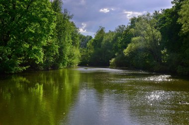Etrafı güzel yeşil bitkilerle çevrili, suya yansıyan küçük bir göl. Fırtınalı bulutlu gökyüzü, bulutların arasından güneş ışınlarını aşıyor. Güzel bir bahar günü.