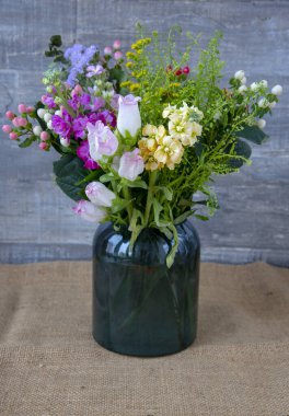 Çiçeklerle dolu bir vazo. Gri duvarın önünde duruyor kaba bir çuval bezinin üstünde.