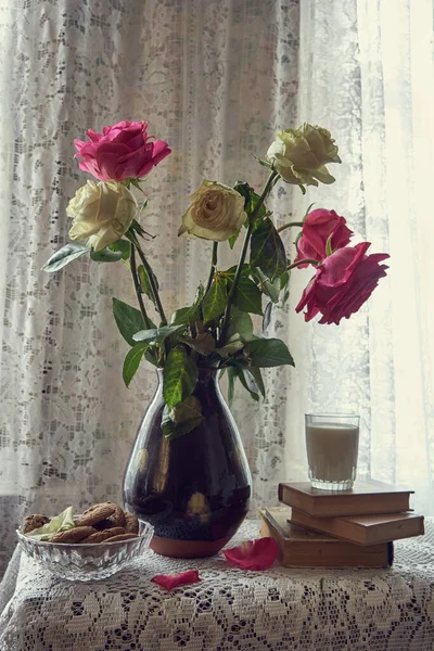 Um buquê de rosas num vaso de cerâmica. Ao lado dele estão livros, um copo de leite e uma tigela de biscoitos. . — Fotografia de Stock