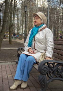 70 'li yaşlarında güzel bir kadın bir park bankında oturuyor. Zarif giyimli ve flörtlü bir şapka takıyor..