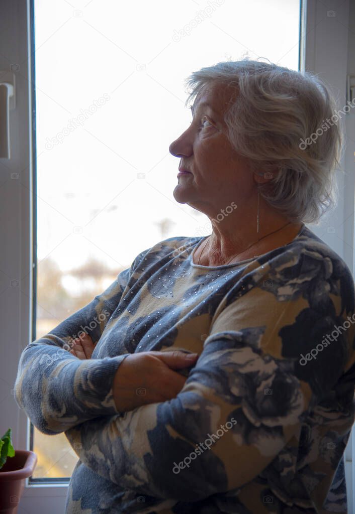A gray-haired elderly woman stands at the window with her arms crossed over her chest. Looks sadly into the distance.