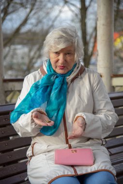Üzgün, yaşlı bir kadın boş bir çanta ve elinde bozuk parayla bir bankta oturuyor..