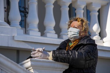 Lastik eldivenler ve koruyucu bir maske takmış yaşlı bir kadın verandada duruyor beyaz taş sütunlar ve balyalara karşı..