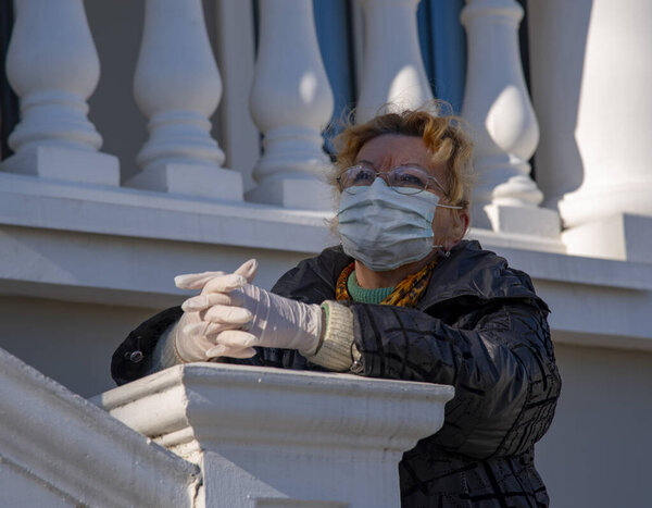 An elderly woman in rubber gloves and a protective mask stands against a backdrop of white stone balustrades.