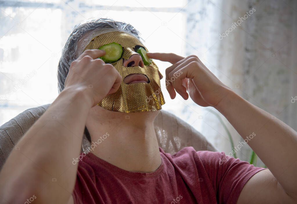 A young man with a gold cosmetic mask on his face and cucumber slices on his eyes. Home Spa, beauty treatments.