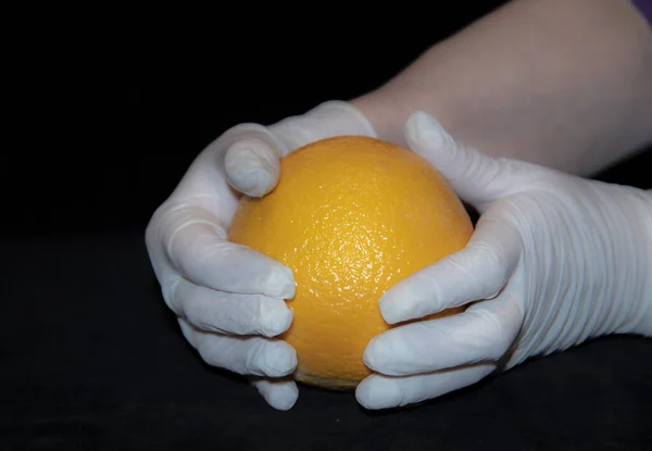 As mãos de uma mulher idosa com luvas de borracha branca seguram uma grande laranja brilhante . — Fotografia de Stock