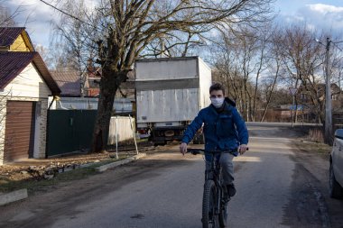 Rusya, Tuchkovo Köyü, Nisan 2020. Koruyucu tıbbi maskeli ve lastik eldivenli genç bir adam köy caddesinde bisiklet sürüyor. Coronavirüs salgını, evde kal..