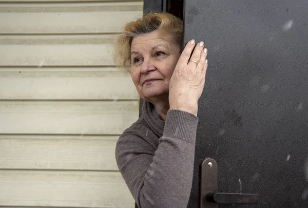 An elegant old woman looks out of a door that is slightly open. Quarantine due to the coronavirus epidemic, stay at home.