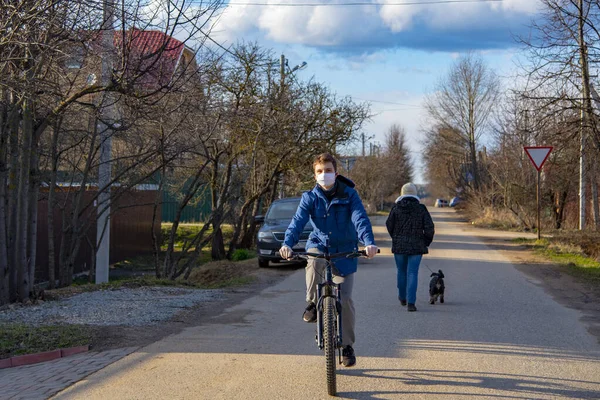 Um jovem com uma máscara médica protetora e luvas de borracha anda de bicicleta ao longo de uma rua da aldeia . — Fotografia de Stock
