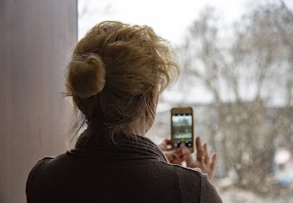 Uma mulher idosa fotografa uma inesperada queda de neve de primavera em seu telefone através de uma porta  . — Fotografia de Stock