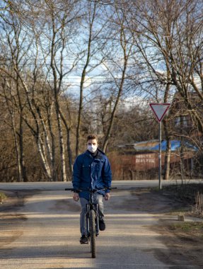 Koruyucu tıbbi maskeli ve lastik eldivenli genç bir adam köy caddesinde bisiklet sürüyor. Coronavirüs salgını, evde kal..