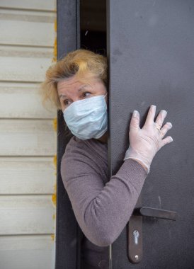 Koruyucu tıbbi maske ve lastik eldiven takmış yaşlı bir kadın kısmen açık bir kapının arkasından dışarı bakıyor. Coronavirüs salgını 2019, evde kalın..