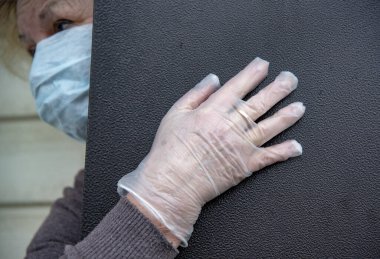 Koruyucu tıbbi maske ve lastik eldiven takmış yaşlı bir kadın kısmen açık bir kapının arkasından dışarı bakıyor. Coronavirüs salgını 2019, evde kalın..