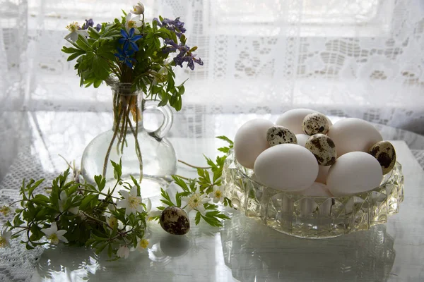 Delicate light Easter still life. Forest primroses in a retro glass bottle and a crystal vase with eggs on a glass surface against a background of lace curtains.