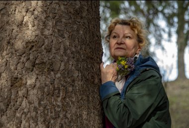 Bir buket bahar ormanı çiçeğiyle zarif, yaşlı bir kadın bir ağaç gövdesine yaslanmış duruyor ve uzaklığa bakıyor..