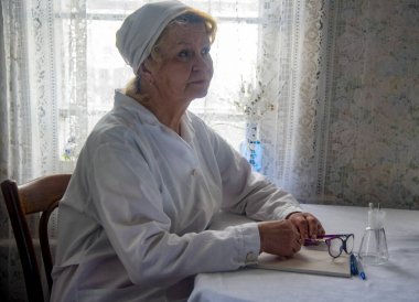 Yaşlı bir kadın doktor hastaların resepsiyonunda bir masaya oturur..