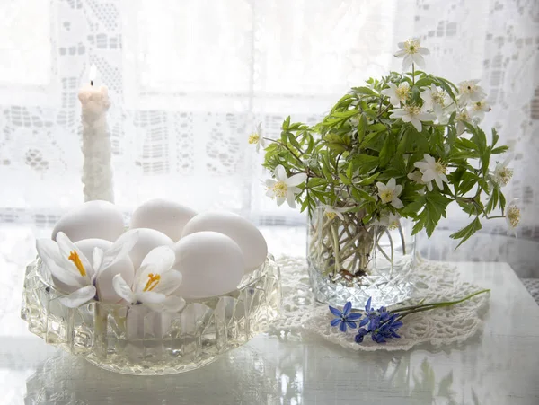 Delicate Easter still life in white tones. Spring flowers in a glass and white eggs in a crystal vase on a white background.