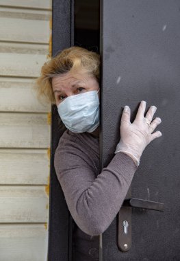 Koruyucu tıbbi maske ve lastik eldiven takmış yaşlı bir kadın kısmen açık bir kapının arkasından dışarı bakıyor. Coronavirüs salgını 2019, evde kalın..