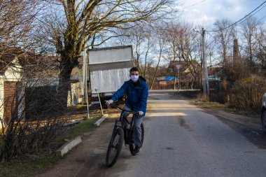 Rusya, Tuchkovo Köyü, Nisan 2020. Koruyucu tıbbi maskeli ve lastik eldivenli genç bir adam köy caddesinde bisiklet sürüyor. Coronavirüs salgını, evde kal..