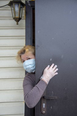 Koruyucu tıbbi maske ve lastik eldiven takmış yaşlı bir kadın kısmen açık bir kapının arkasından dışarı bakıyor. Coronavirüs salgını 2019, evde kalın..