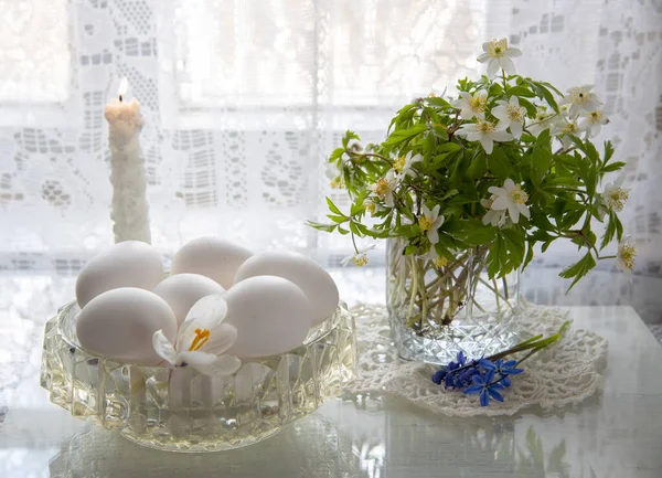 Delicate Easter still life in white tones. Spring flowers in a glass and white eggs in a crystal vase on a white background.