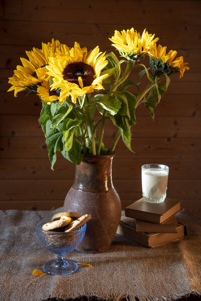Букет ярких подсолнухов в глиняном кувшине, книги, стакан молока и миска печенья на грубой скатерти на деревянном фоне
.