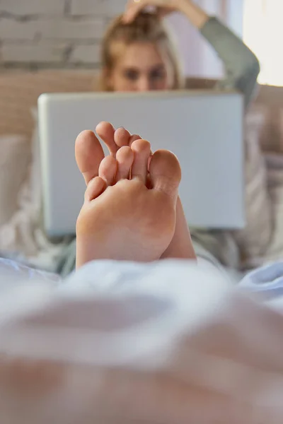 Focus on the feet in the foreground. A teenage girl is lying on a bed with a laptop.