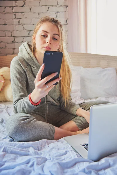 Una chica rubia está sentada en la cama en un chándal gris con un teléfono y un portátil . — Foto de Stock