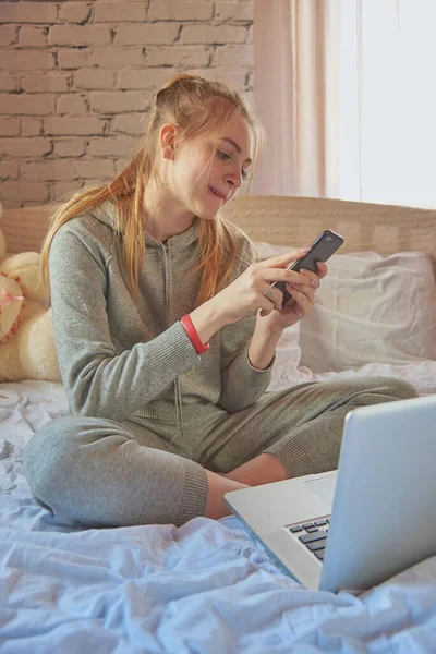 Una chica rubia está sentada en la cama en un chándal gris con un teléfono y un portátil . — Foto de Stock