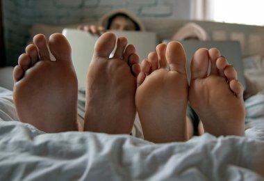 Ön planda çıplak ayaklar. İki genç dizüstü bilgisayarlarla bir yatakta yatıyor..