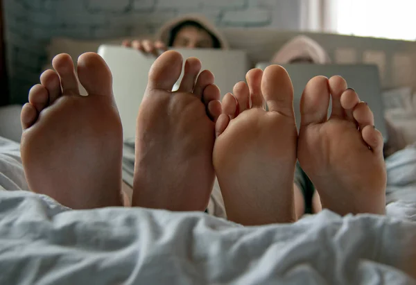 Bosé nohy v popředí. Dva teenageři leží na posteli s laptopy. — Stock fotografie