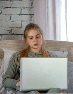 Sarışın bir kız gri eşofmanlı ve dizüstü bilgisayarlı bir şekilde yatakta oturuyor. Mesafe öğrenme, online alışveriş, iletişim.
