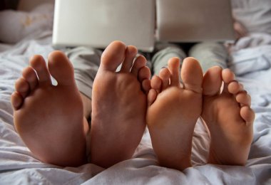 Ön planda çıplak ayaklar. İki genç dizüstü bilgisayarlarla bir yatakta yatıyor. Sosyal ağlarda uzaklık öğrenimi, iletişim.